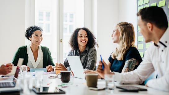 Un groupe d’employé(e)s de bureau assis et discutant ensemble autour d’un grand bureau lors d’une réunion matinale.