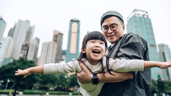 Happy little Asian girl having fun with her father outdoors in urban park. She is pretending to be an airplane, flying up in the sky while her father lifting her into the air. Family love and lifestyle