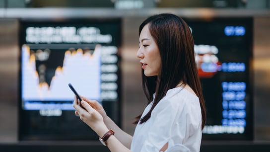Young Asian businesswoman using smartphone against stock exchange market display screen board in downtown financial district