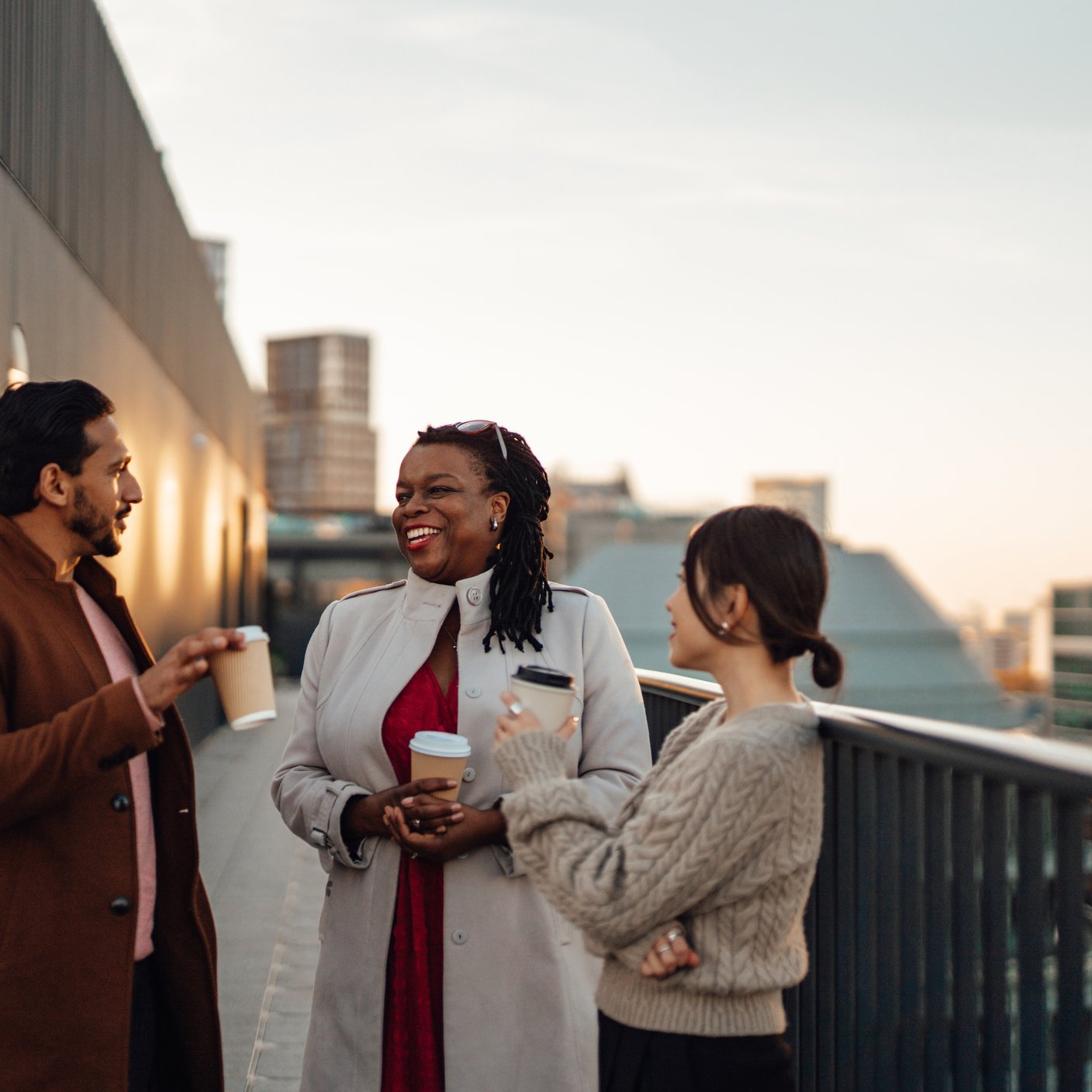 Cheerful multi-ethnic business people standing by railing at rooftop during coffee break. Talking about work and life together.