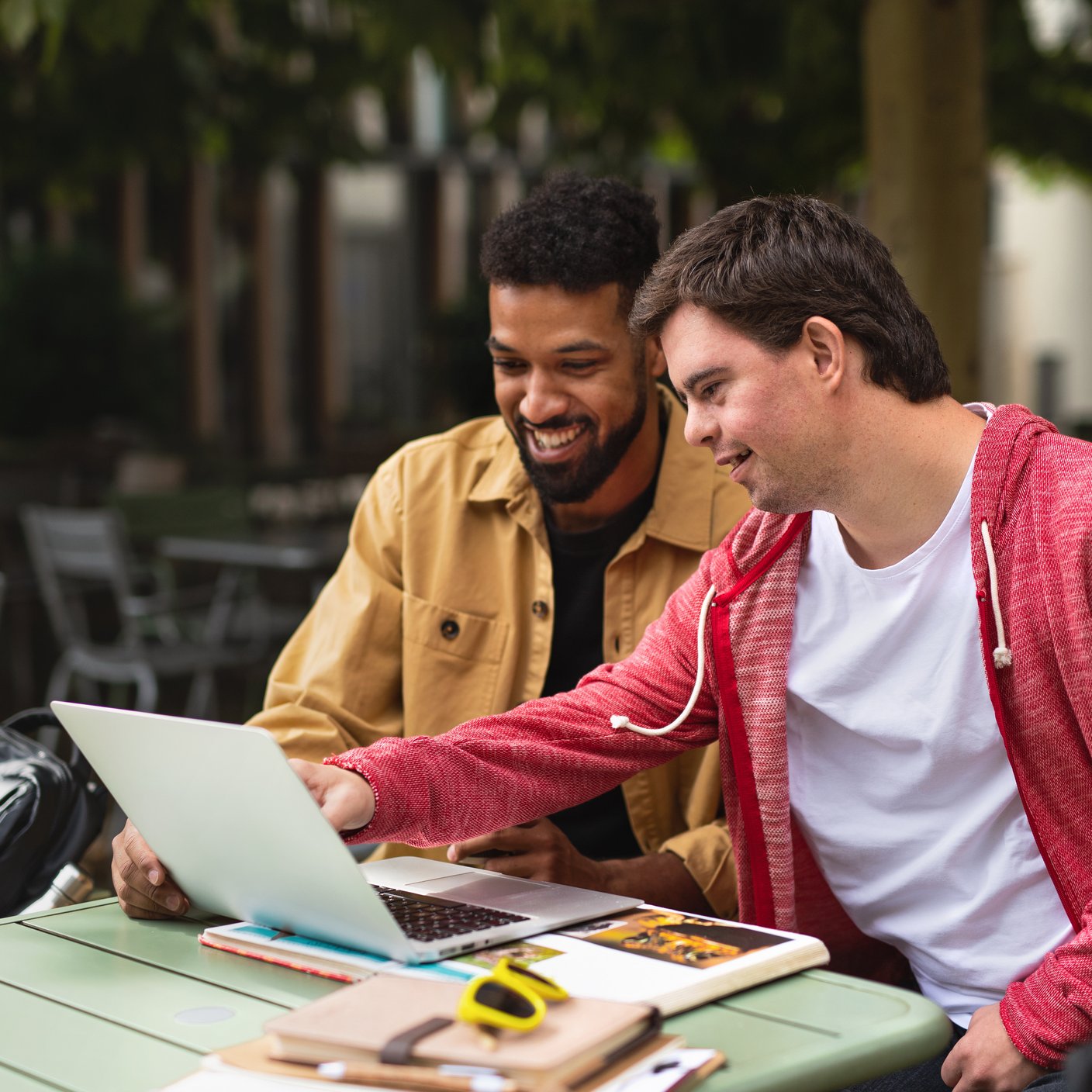 A young man with Down syndrome with his mentoring friend sitting outdoors using laptop