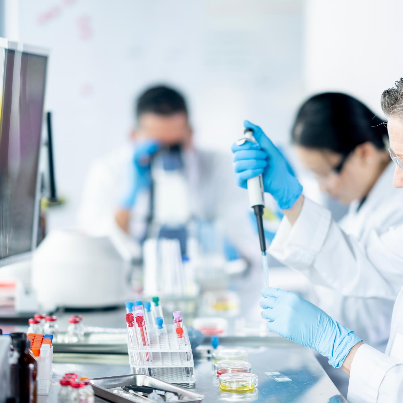 A small team of adults work together in a Medical Lab as they draw samples and take a closer look under microscopes. They are each dressed professionally in lab coats and are wearing protective eyewear as they analyze the data.