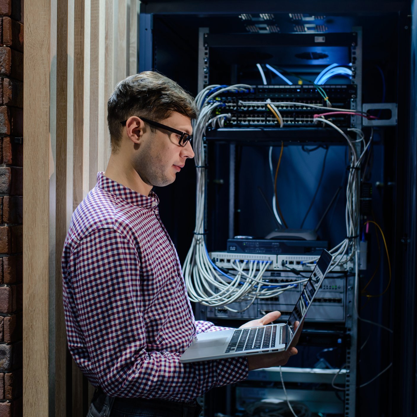 In the server room a technician/ It engineer/ with glasses hold the notebook and adjust the internet connecting or fix the server error.