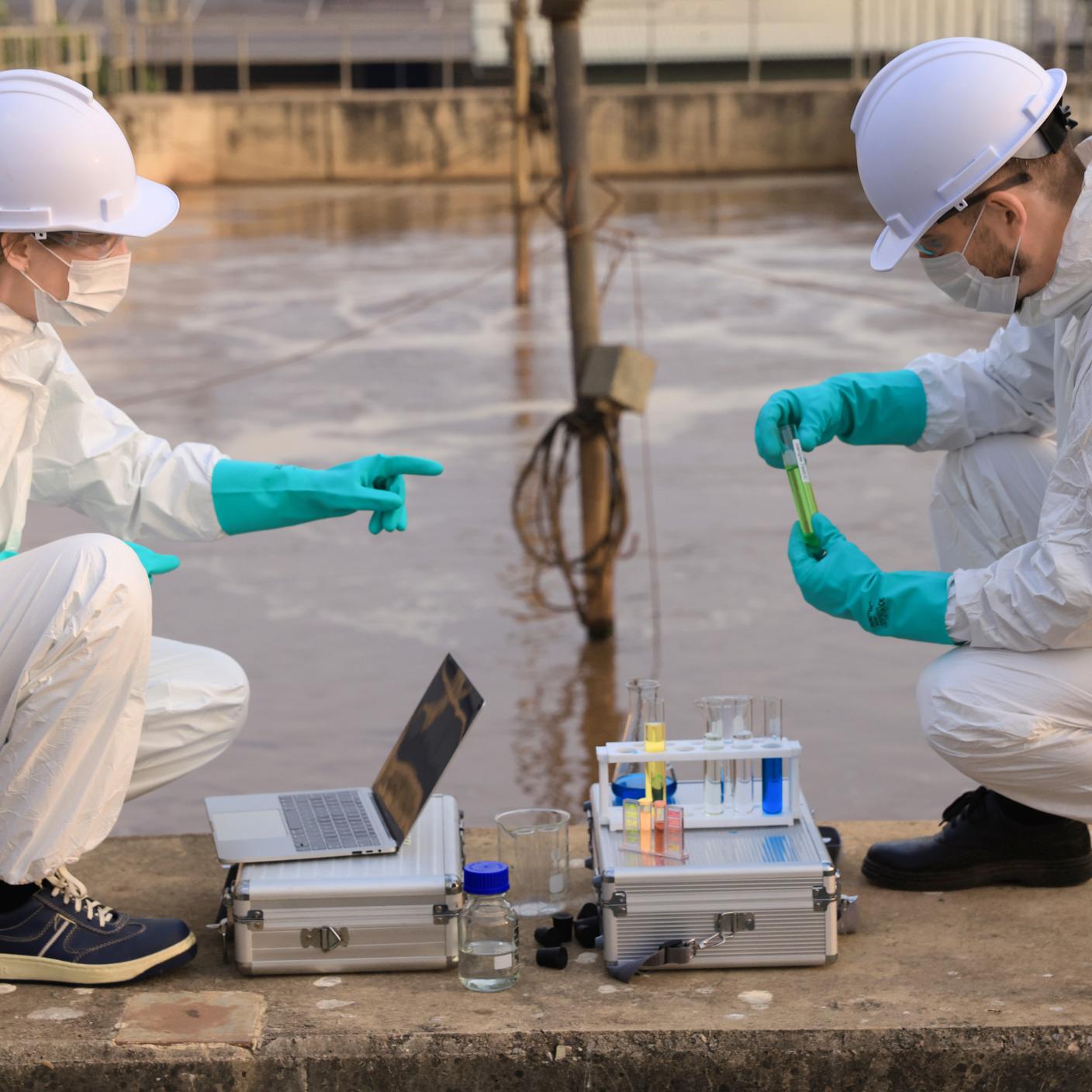 Two environmental technicians in protective gear collect and analyze water samples using portable testing equipment.