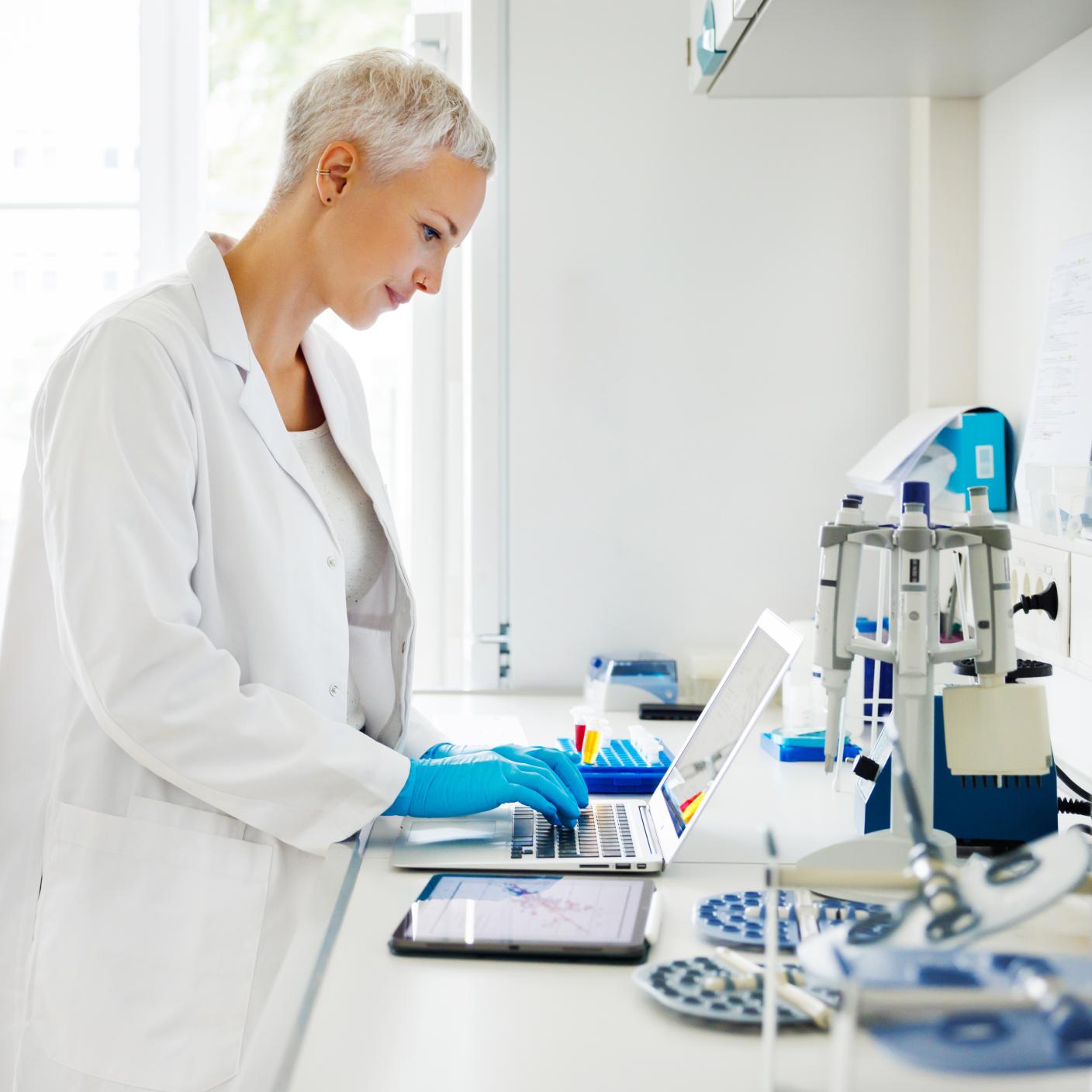 A scientist, dressed in a lab coat and gloves, focuses intently on analysing data using a laptop and tablet. 