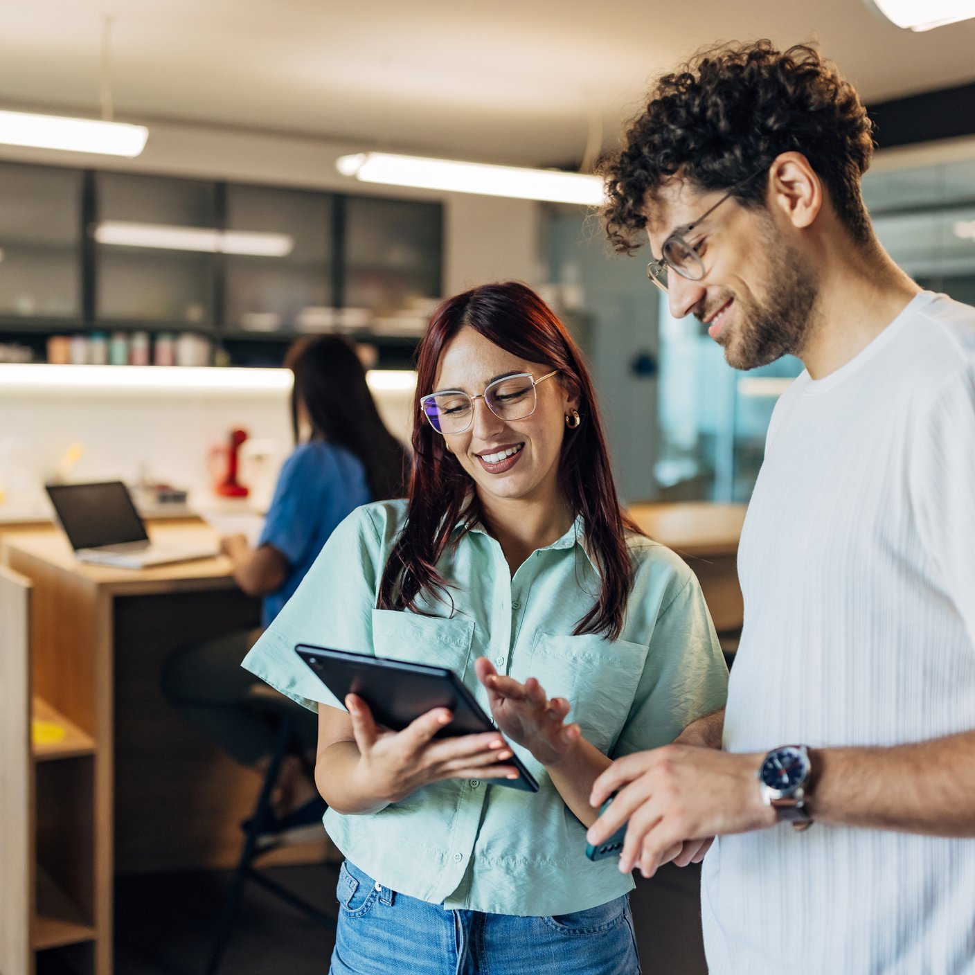 Dos compañeros de trabajo sonrientes miran la pantalla de una tableta digital mientras están de pie en un espacio de trabajo híbrido.