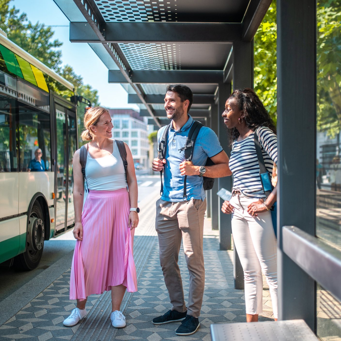 A group of diverse friends, men and women, stand engaging in conversation at a sunny urban bus station, dressed in casual attire, anticipating their commute.