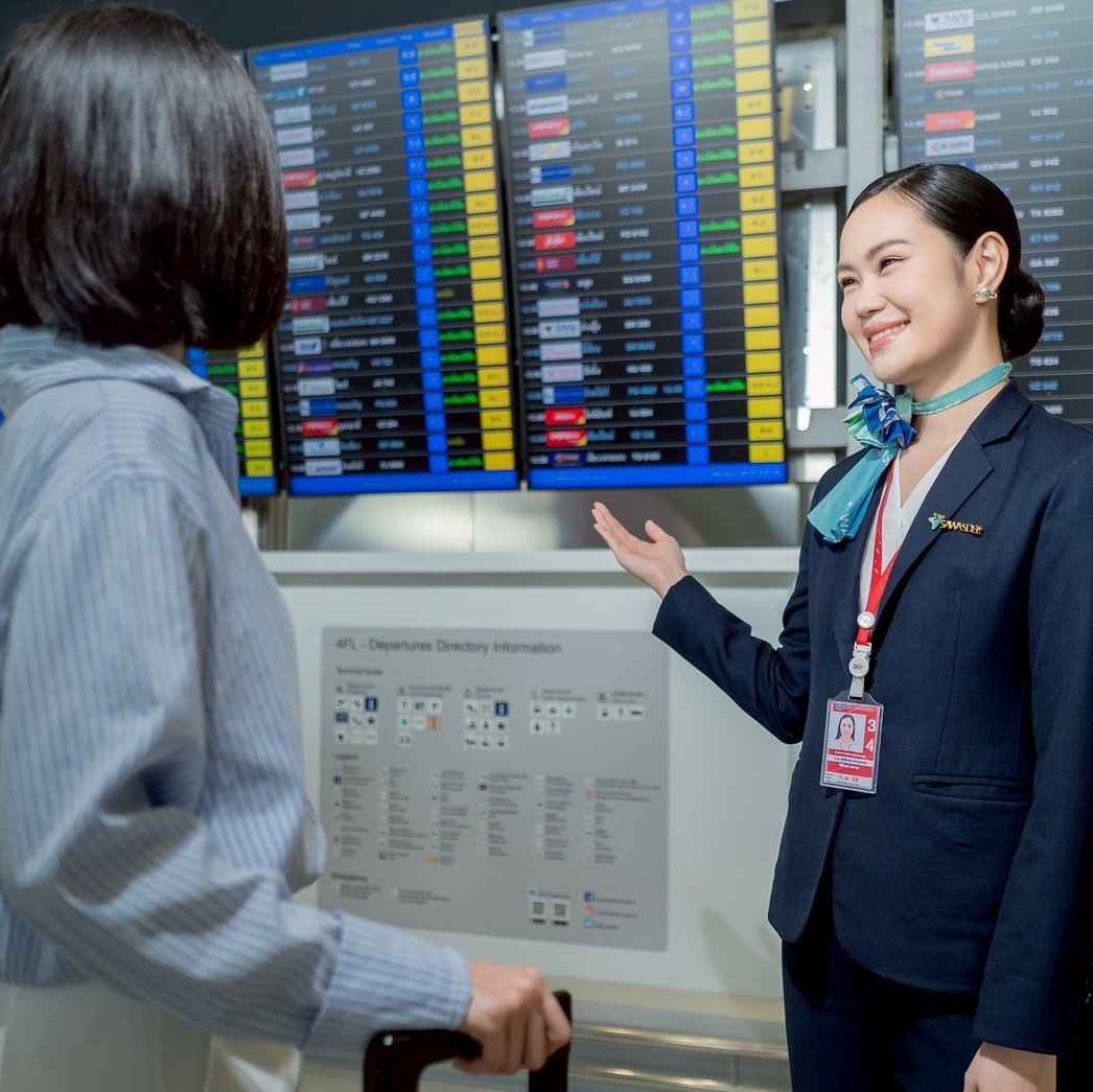 A female airport customer service agent in a dark navy uniform and teal neck scarf smiles while directing a passenger in front of a large departures information board.