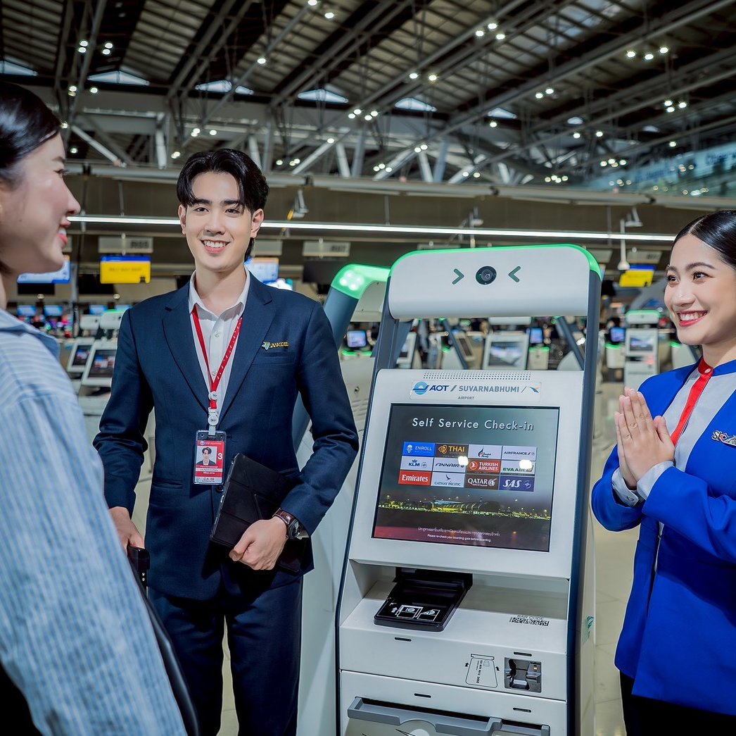 Two airport staff members, a man in a dark suit and a woman in a blue uniform, greet a passenger at an AOT Suvarnabhumi Airport self-service check-in kiosk.
