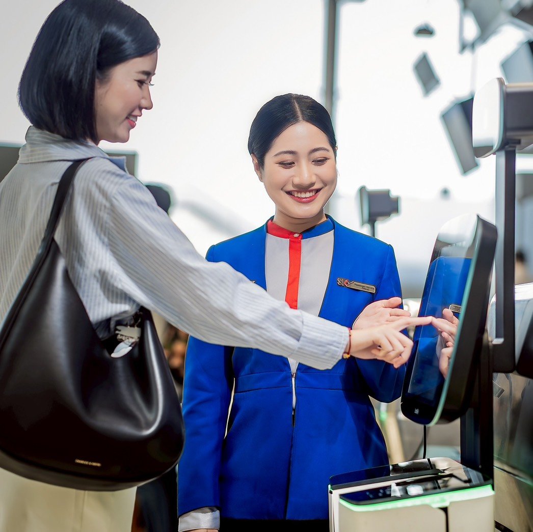 A female airport staff member in a blue uniform smiles as she guides a passenger through the self-check-in process at a touchscreen kiosk. 