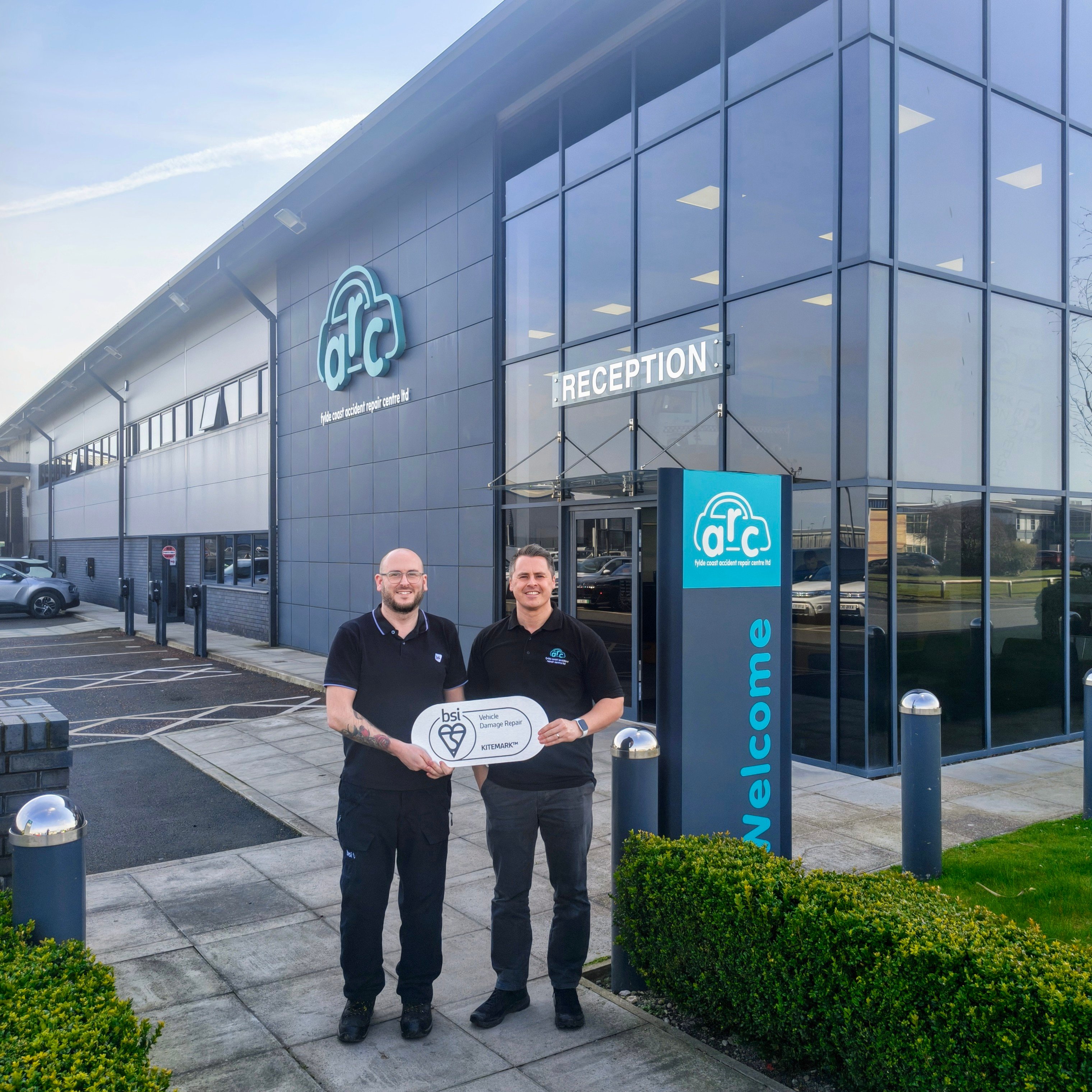 Two ARC team members in branded black polo shirts stand outside the company's reception entrance, holding a large BSI Kitemark for Vehicle Damage Repair certification plaque. 