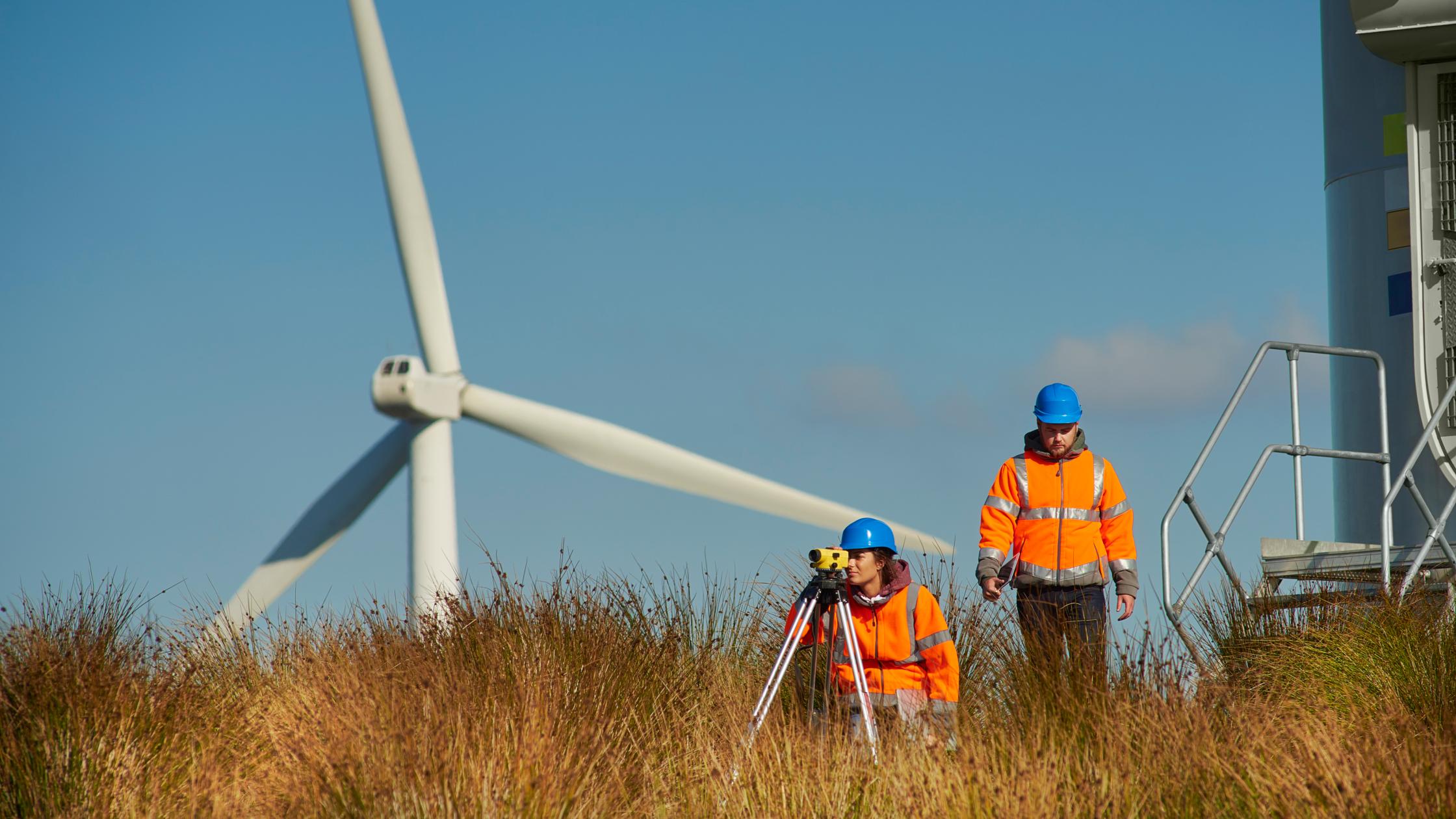 two wind farm engineers leaving the control panel of a wind turbine. They are assessing their readings on a digital tablet as they walk away. In the foreground a theodolite can be seen.