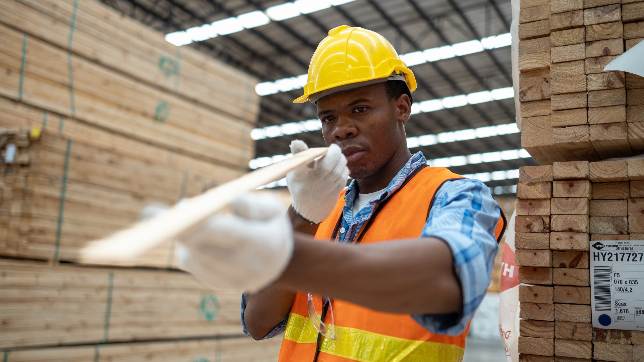 A diligent African worker in a vibrant safety vest and helmet examines a piece of lumber with precision.